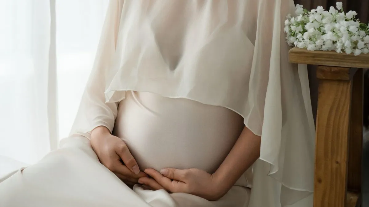 Femme enceinte assise près d'une fenêtre, les mains sur le ventre.
